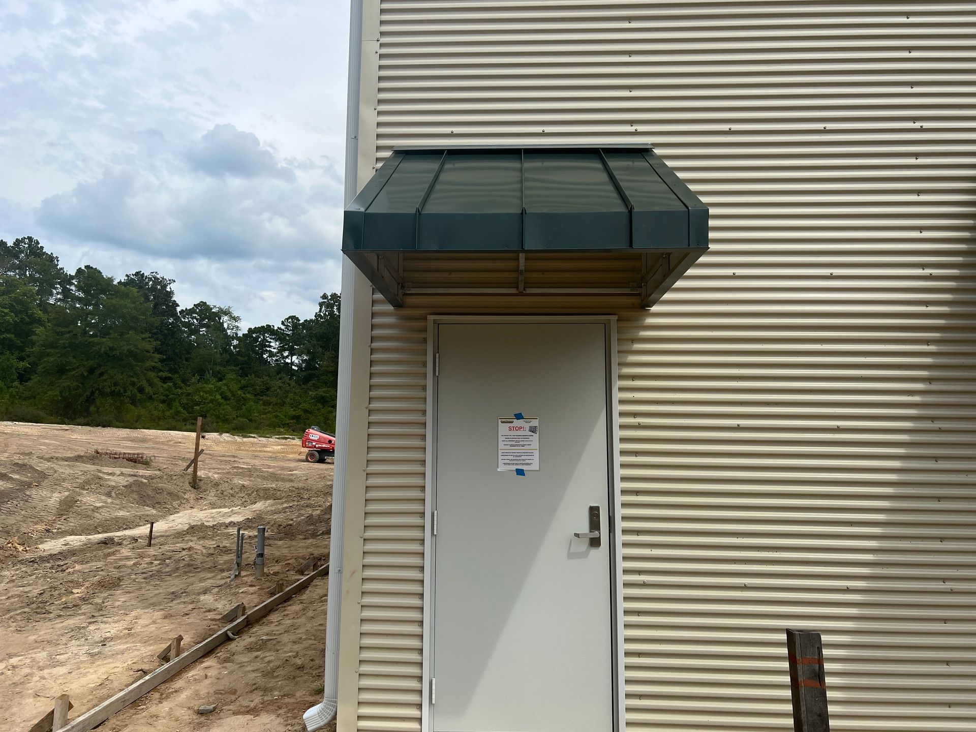 White utility door with awning on beige corrugated building beside a dirt lot and trees