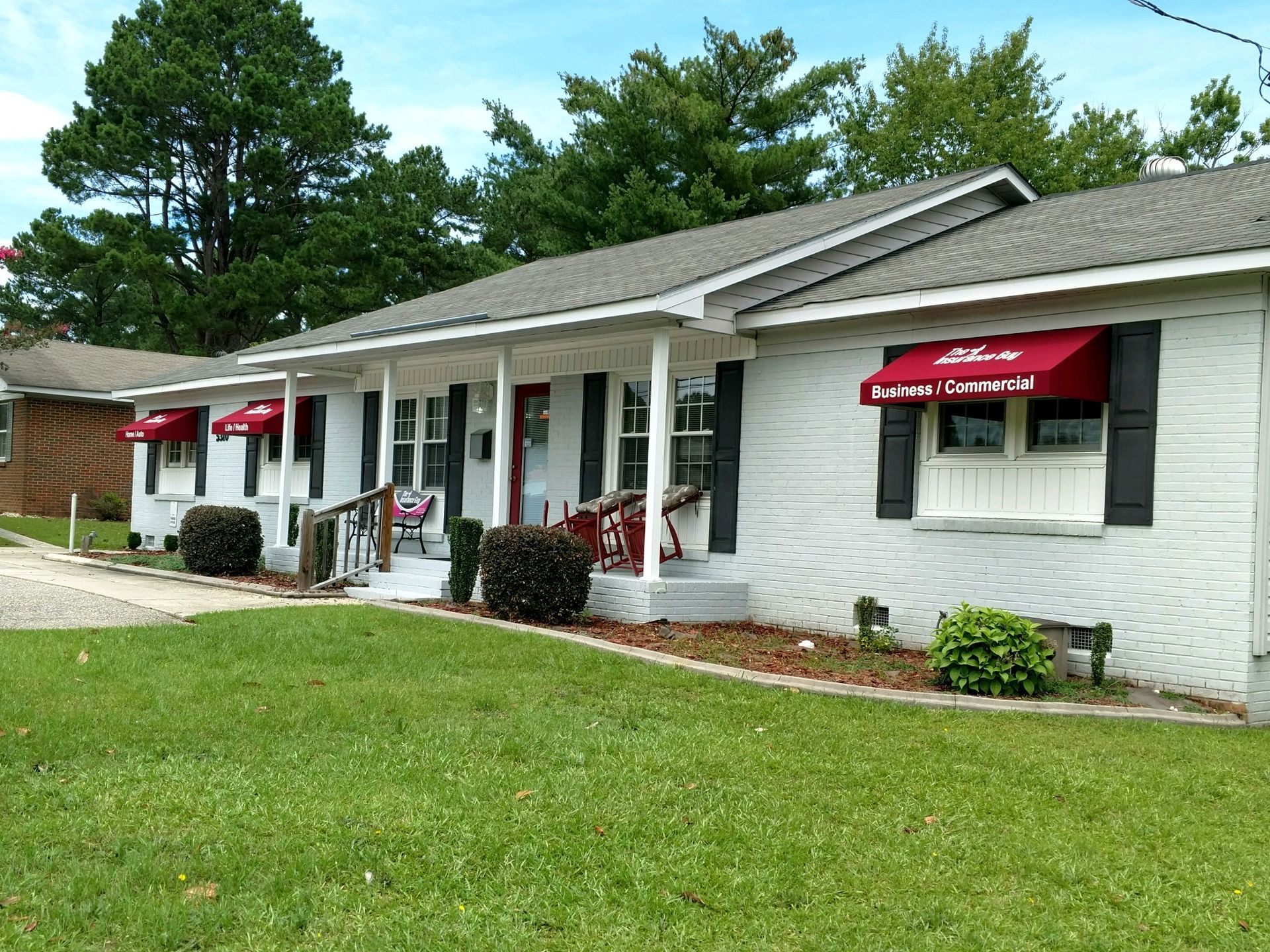 White storefront with red awnings and a covered porch beside a green lawn and trees