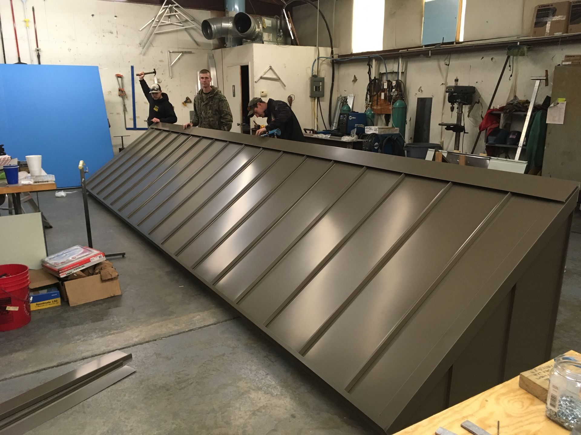 Large brown metal roof panel with standing seams in a workshop, with workers in the background