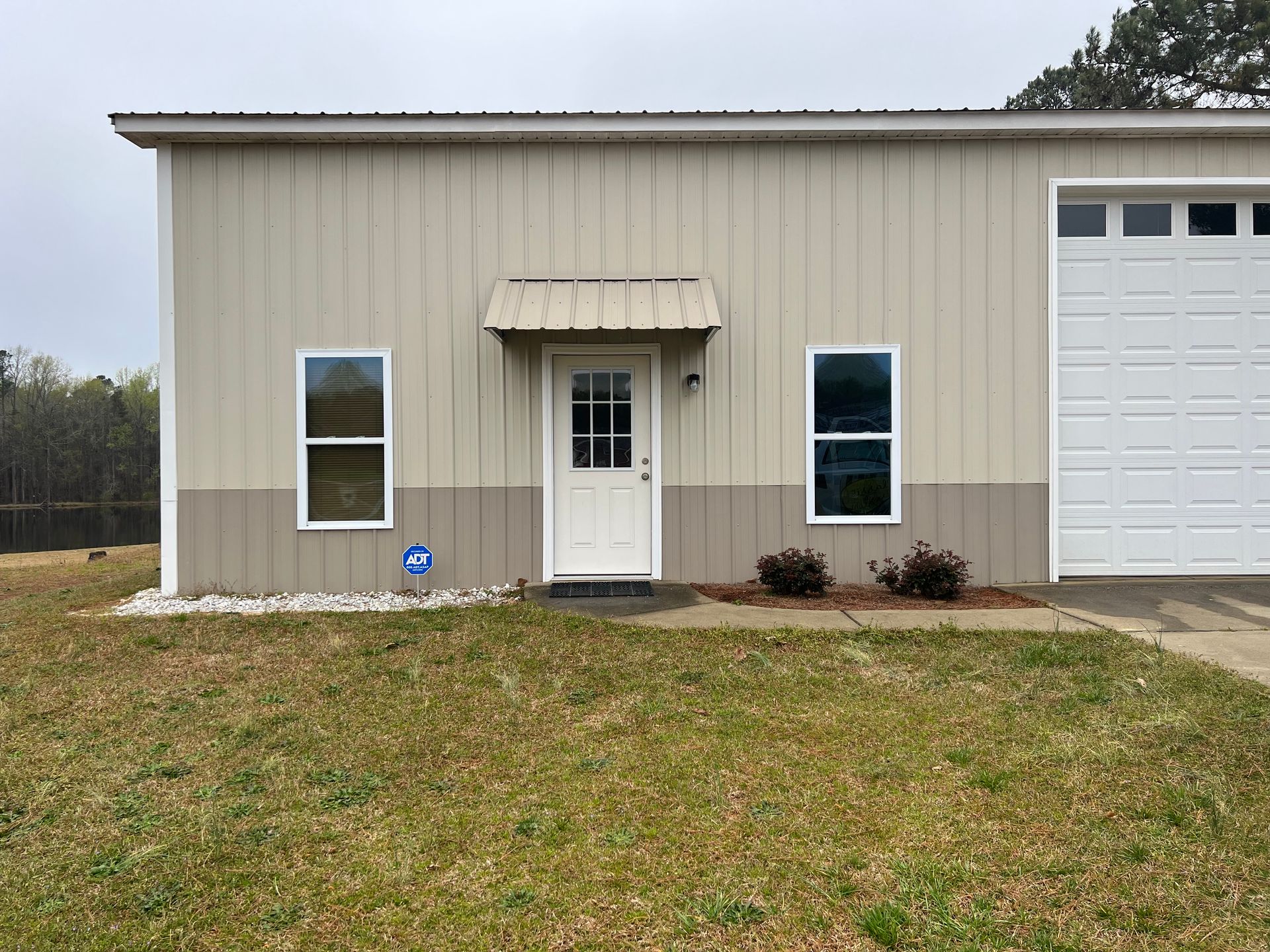 Small beige building with a white door, two windows, and an attached garage on a grassy lot