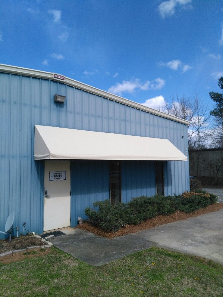 Light blue building entrance with white awning, shrubs, and sidewalk under a blue sky.