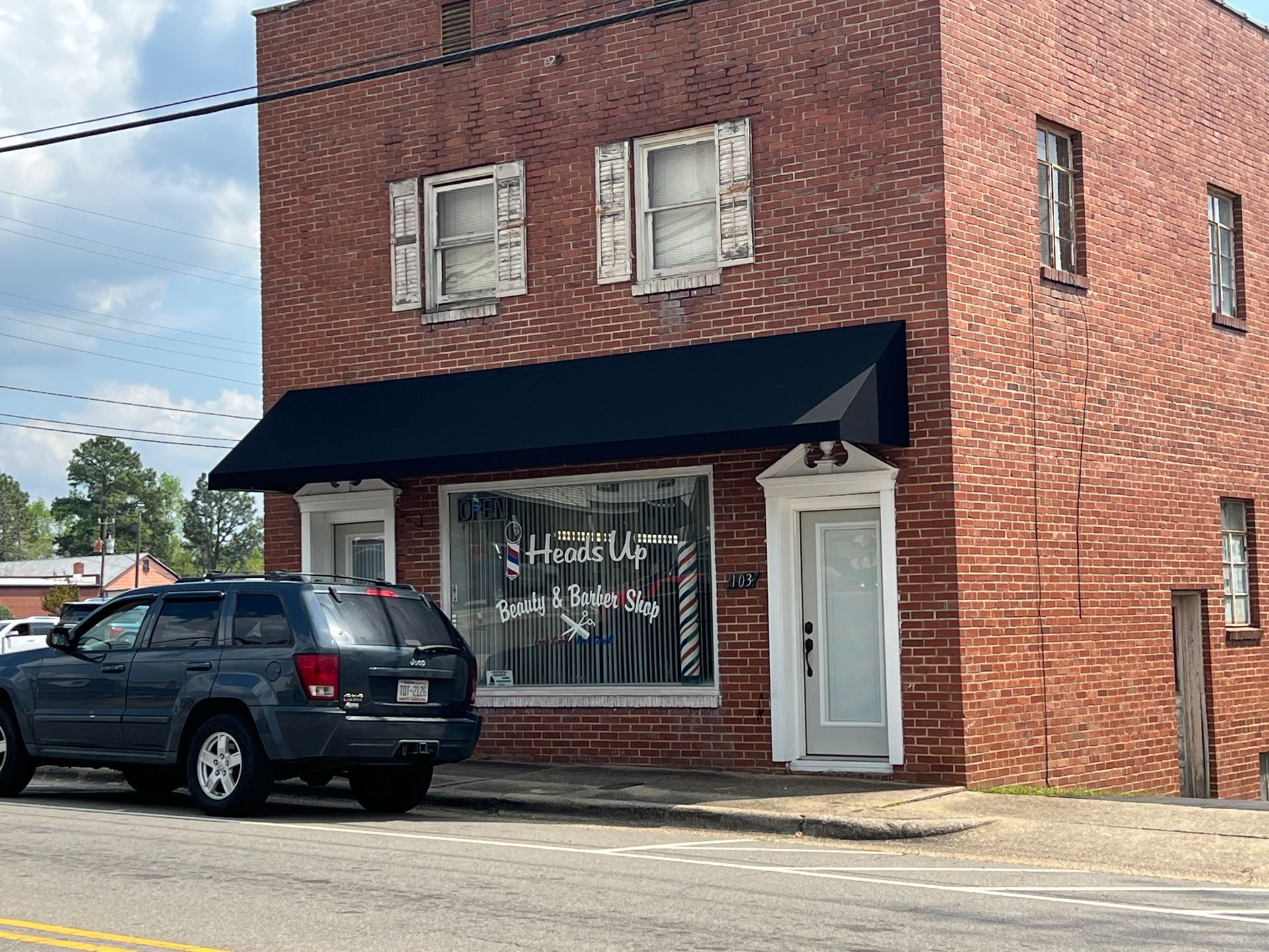Brick storefront with black awning, white door, and parked black SUV on a street corner