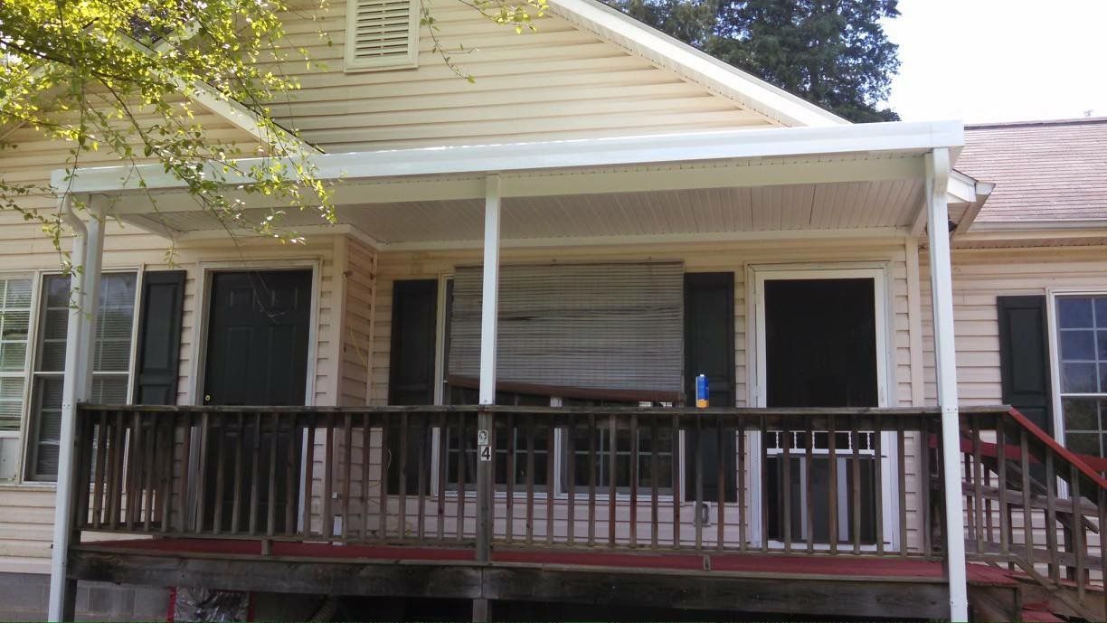 Front porch of a yellow house with dark shutters and a wooden railing, viewed from the yard