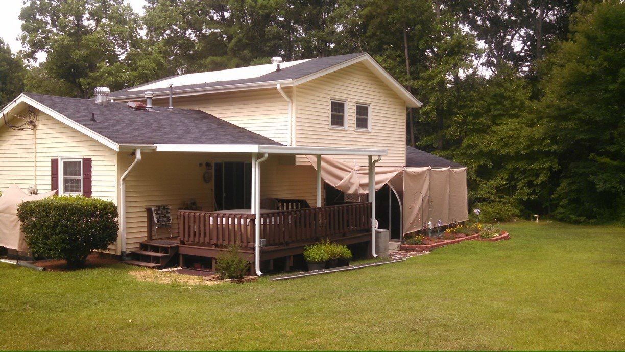 House with tan siding, brown porch, and an attached canopy in a grassy yard beside trees