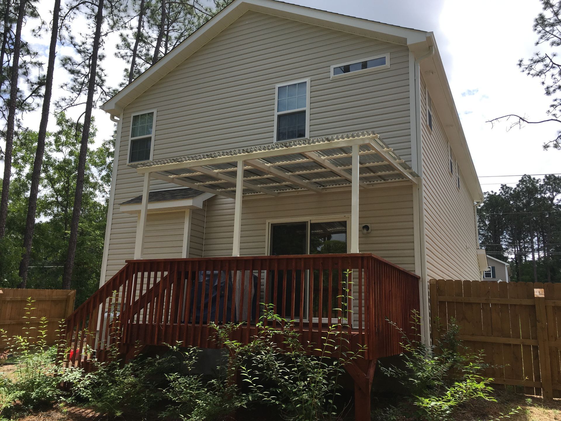 Two-story tan house with a red wooden deck and a partially built upper balcony in a wooded yard