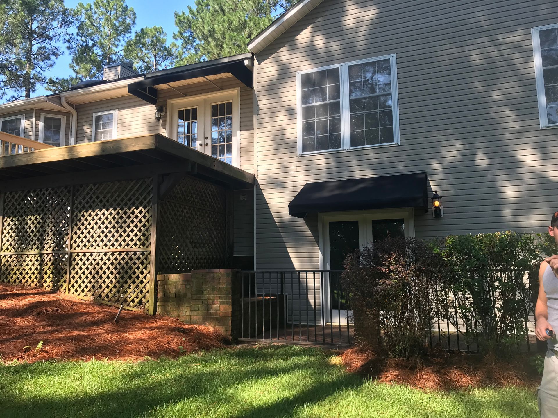 Two-story house exterior with beige siding, black awnings, and a covered side porch in a sunny yard