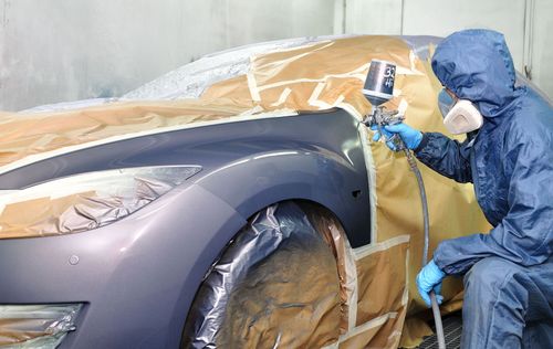 Technician in protective suit spray-painting a car in a covered auto body shop.