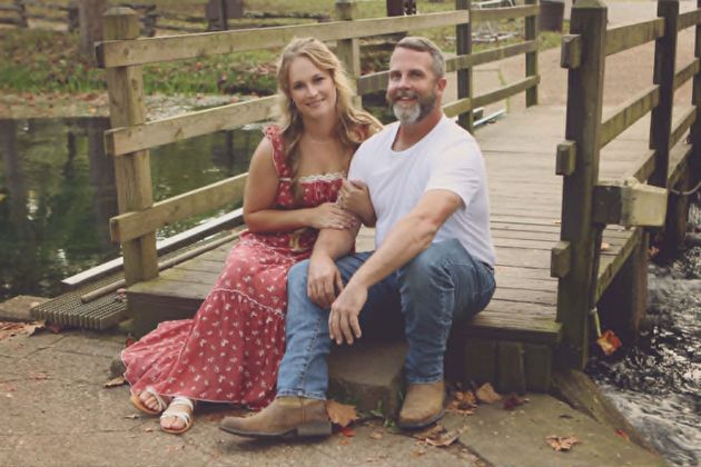 Couple sitting on wooden bridge, smiling. Woman in red dress, man in white shirt and jeans. Outdoors.