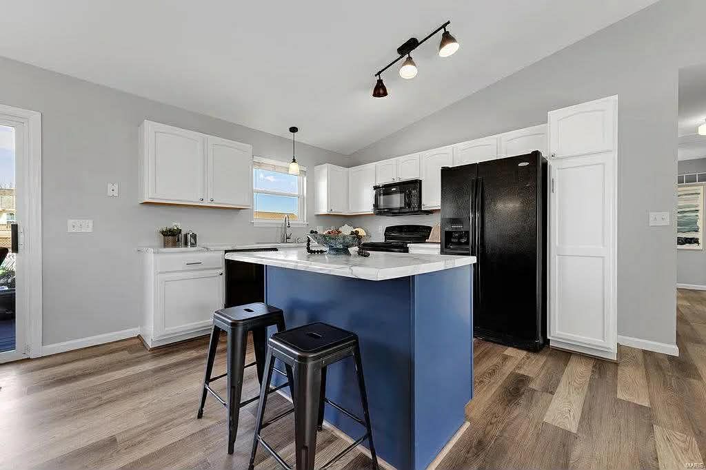 A kitchen with white cabinets, a blue center island with two black stools, a black refrigerator, and light wood floors.