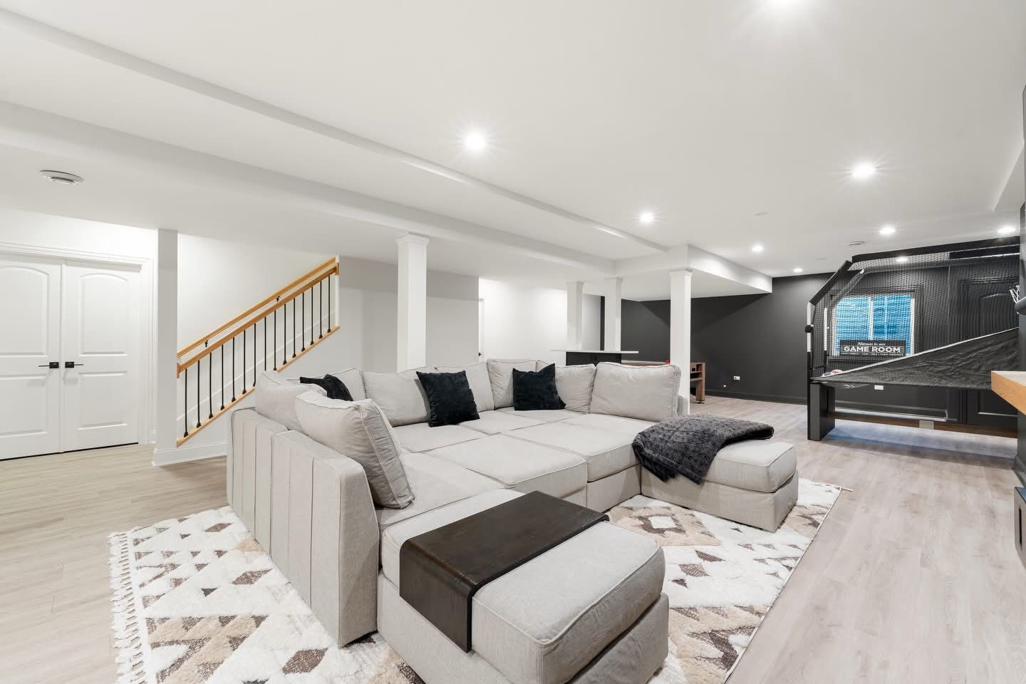 Spacious, modern basement living area with sectional sofa, rug, stairs, and a dark wall with a table.