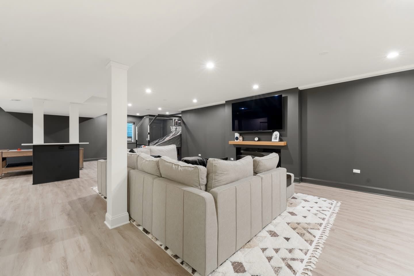 Modern basement living area with sectional sofa, TV, and bar area. Gray and neutral tones.