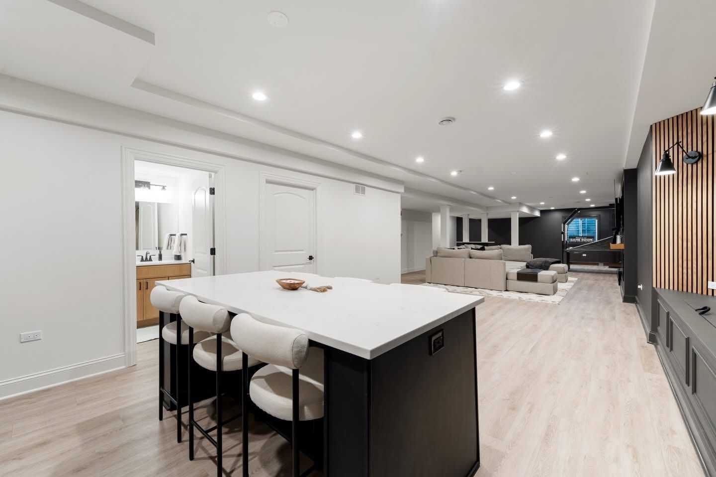 Modern open-plan basement with white kitchen island, seating, and living area with couch, dark accent wall, and recessed lighting.