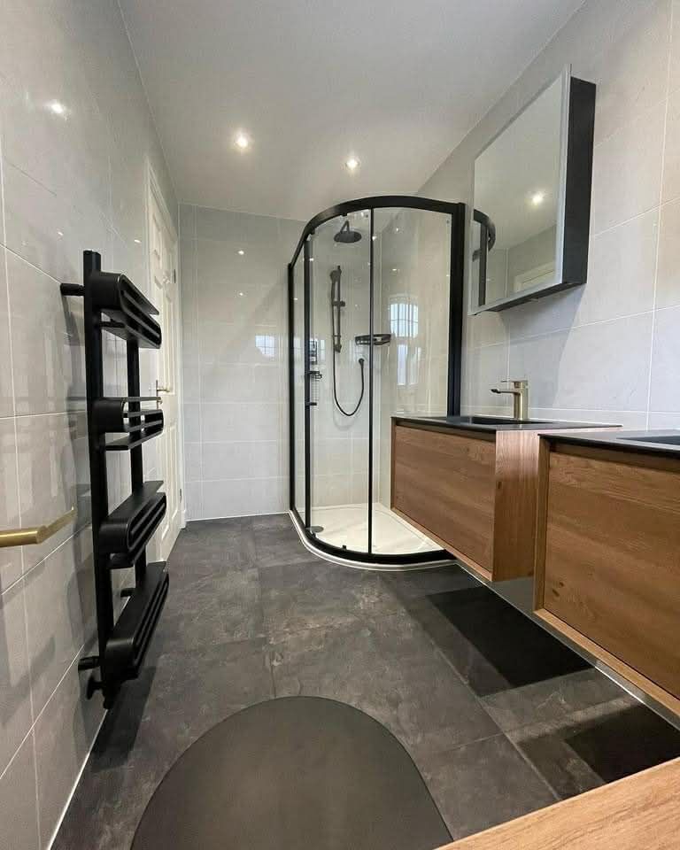 Modern bathroom with black-framed shower, wooden vanity, gray tile, and black towel rack.