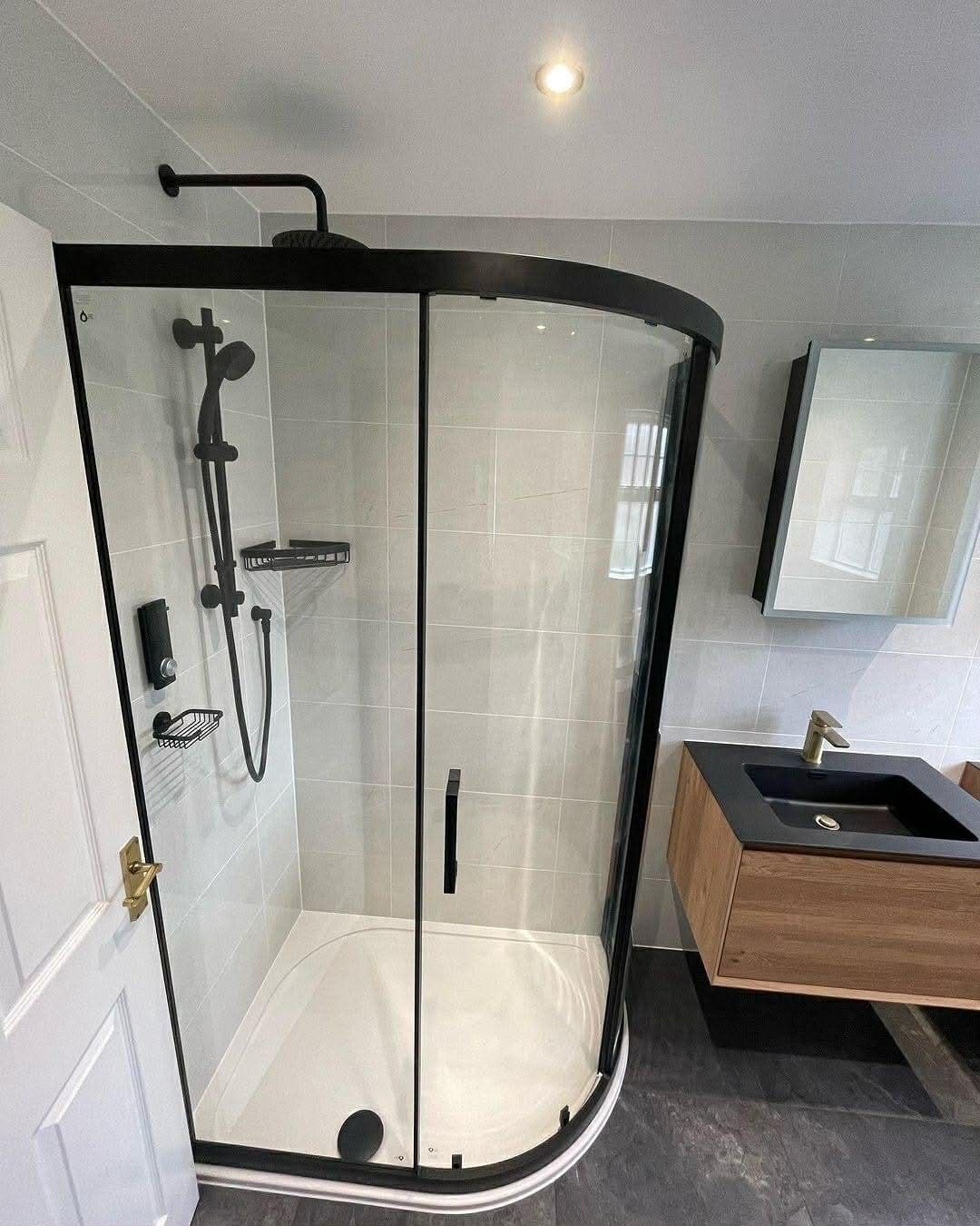 Curved black-framed shower with black fixtures; wooden vanity with a black sink and mirror to the right.