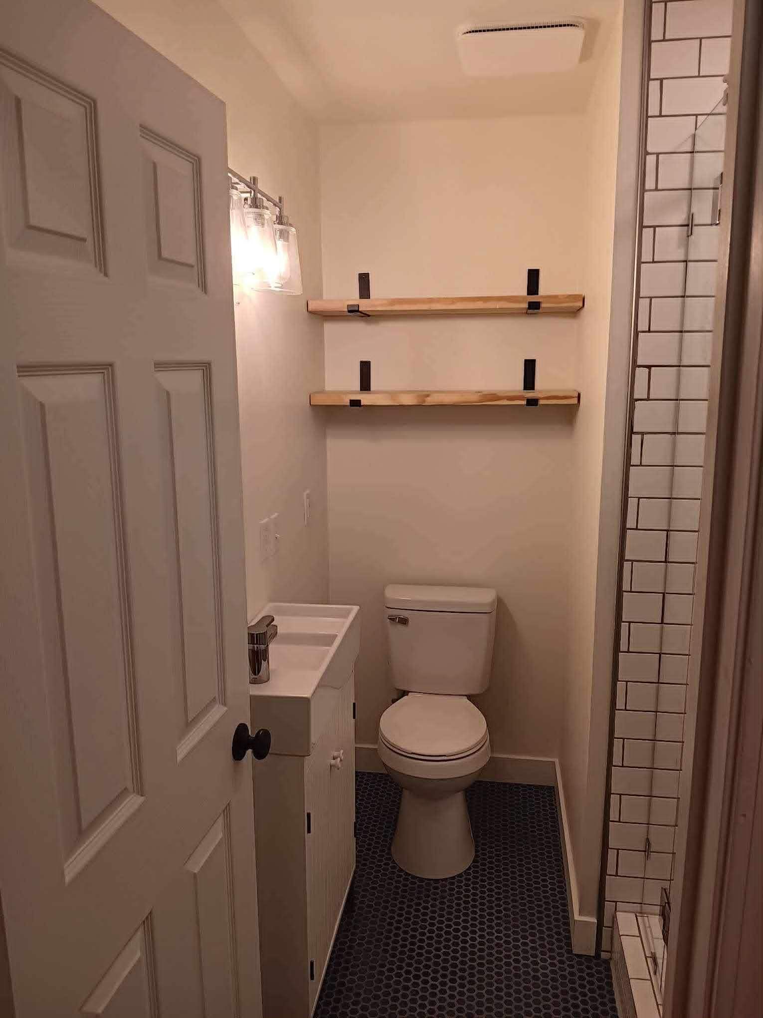 Small white bathroom with toilet, shelves, and a tiled shower; blue and white tiled floor.