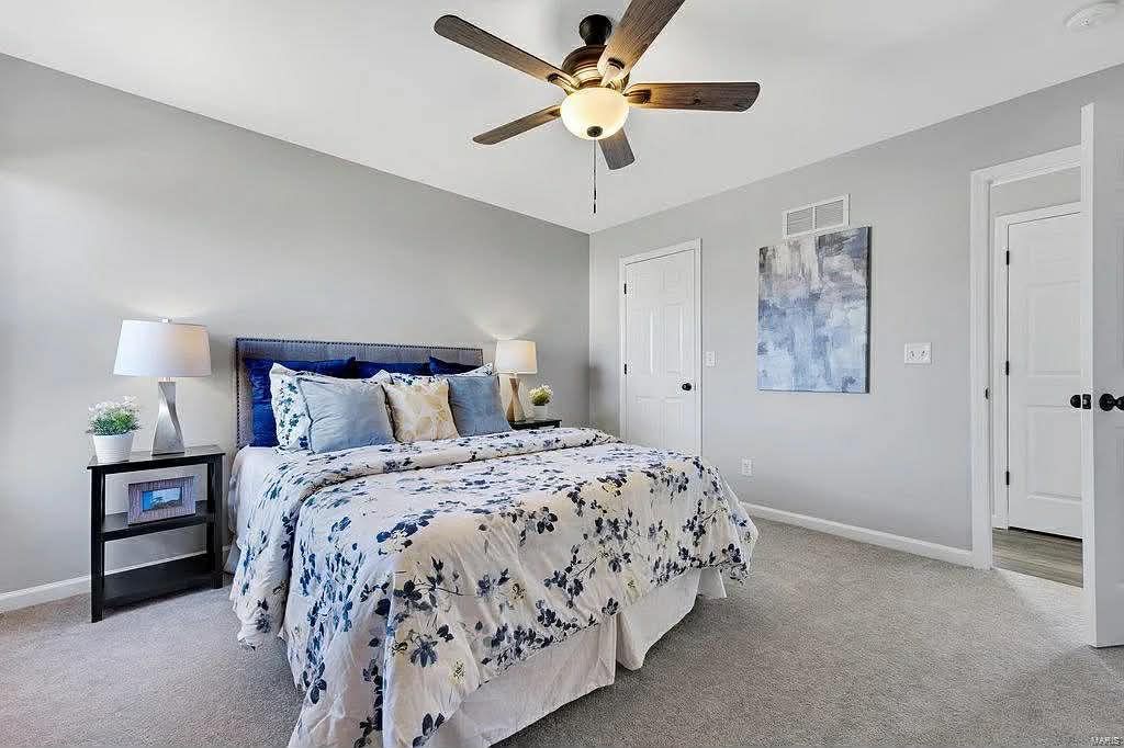 A furnished bedroom with a king-sized bed, floral comforter, gray walls, ceiling fan, and carpeted flooring.