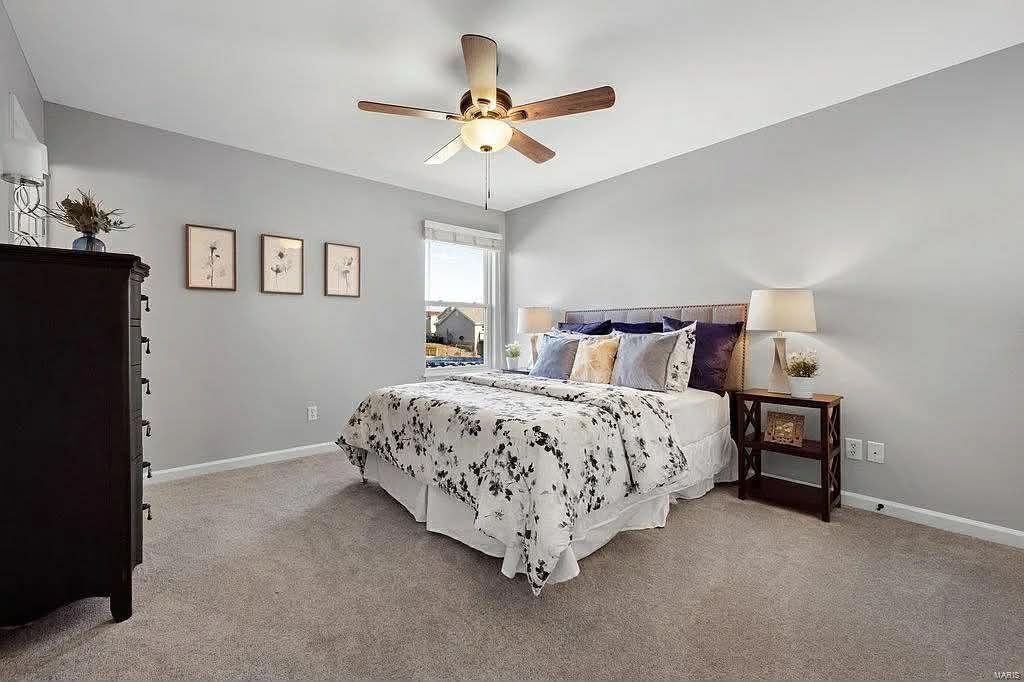A bedroom with a queen bed featuring a floral duvet, a dark wood dresser, a bedside table with a lamp, and a ceiling fan.