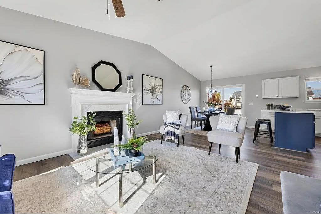 A modern, bright living room features a fireplace, a coffee table on a large area rug, and an open kitchen in the back.