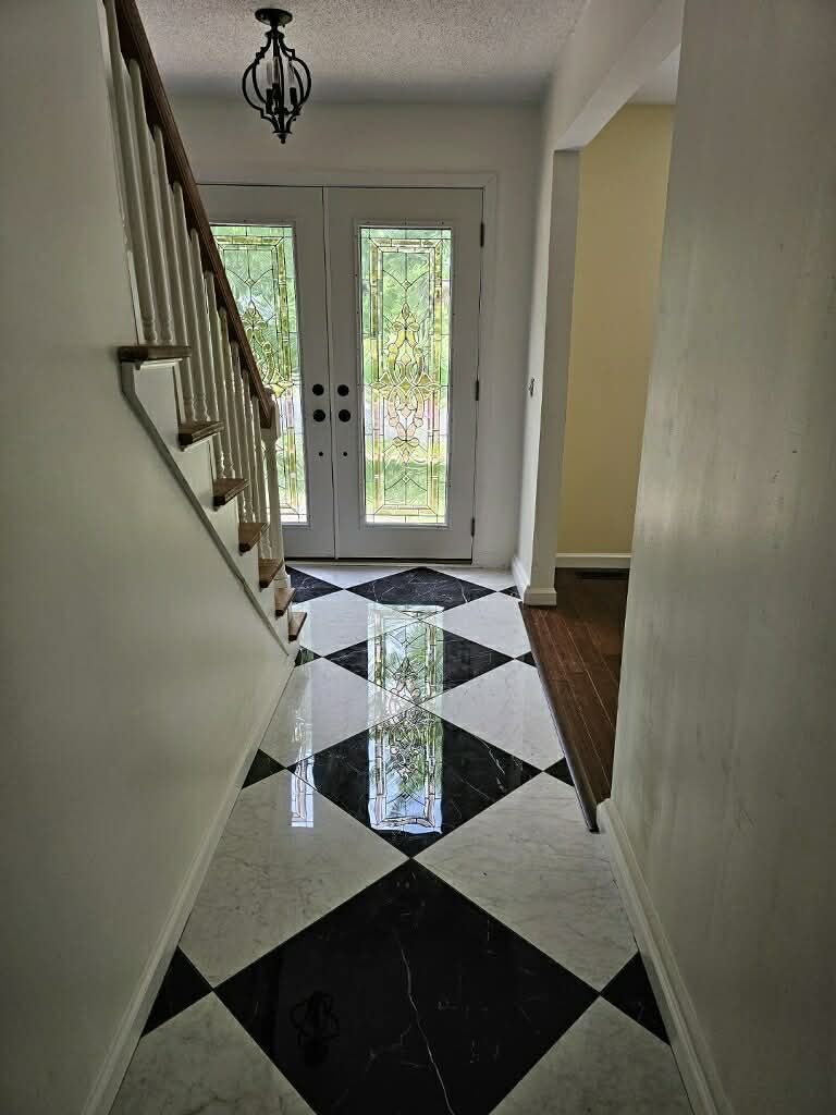 A bright entryway featuring a black-and-white diamond tiled floor, a staircase with a wooden railing, and double doors.