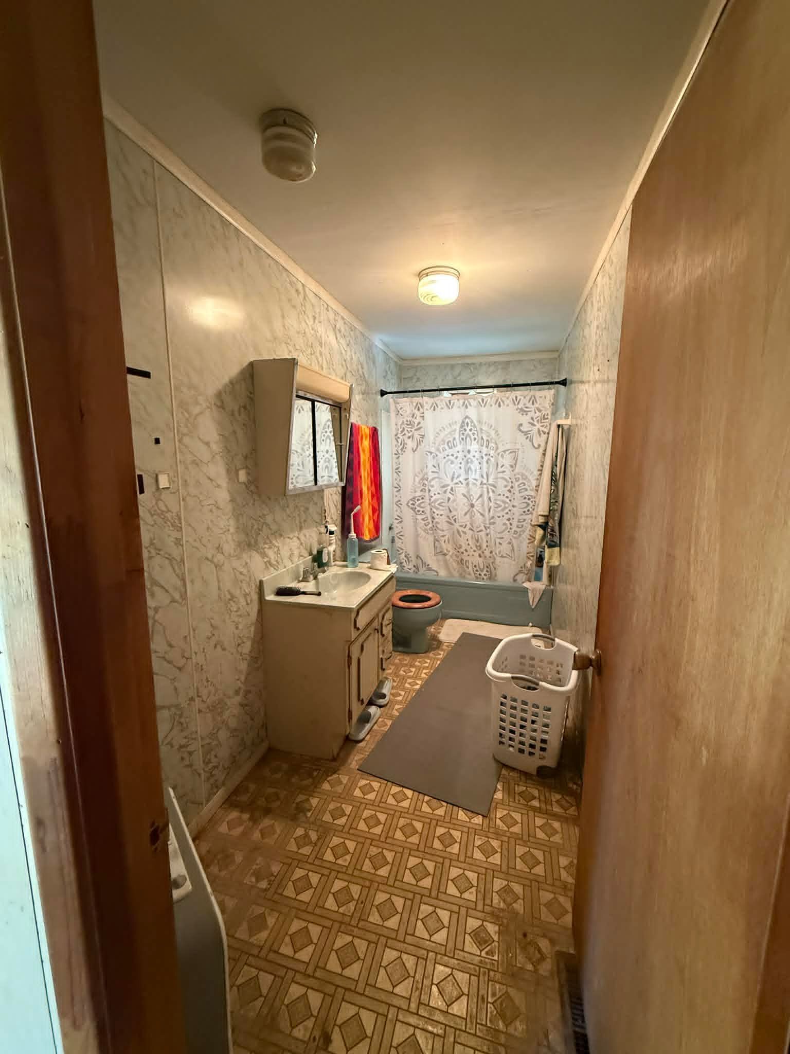 Narrow bathroom with a sink, tub, and laundry hamper. The floor has a patterned design, and the walls are panelled.