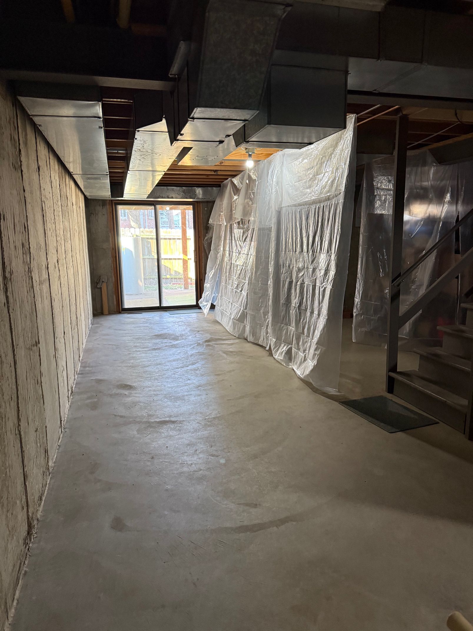 Basement hallway with concrete floor and unfinished walls. Plastic sheeting covers a section. Sliding glass door visible.