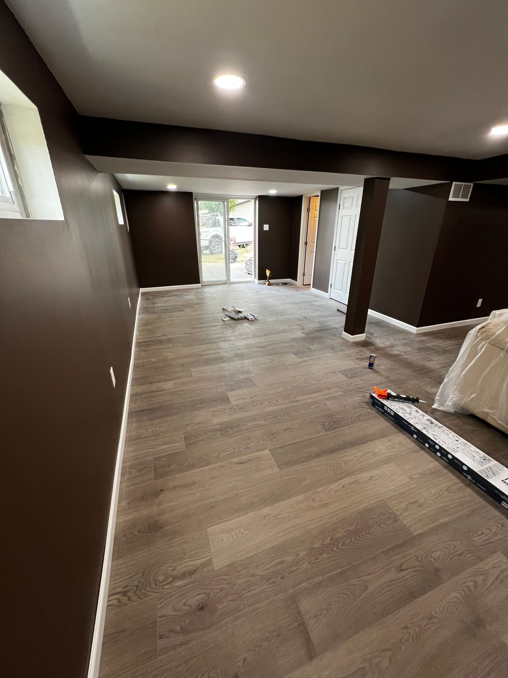 Brown-walled room with wooden floor, sliding glass door, and recessed lighting. Construction materials and tools present.