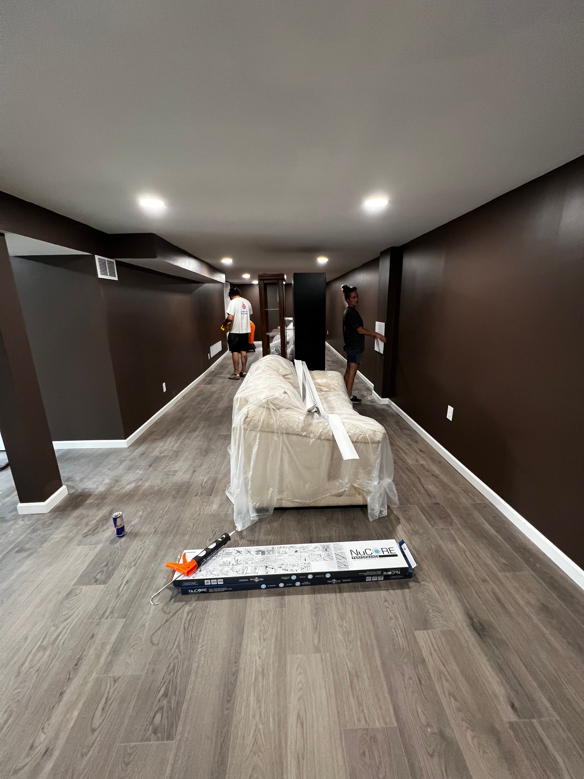 Room with dark brown walls, gray flooring, and white trim. Two people in the distance, working. Couch covered in plastic.
