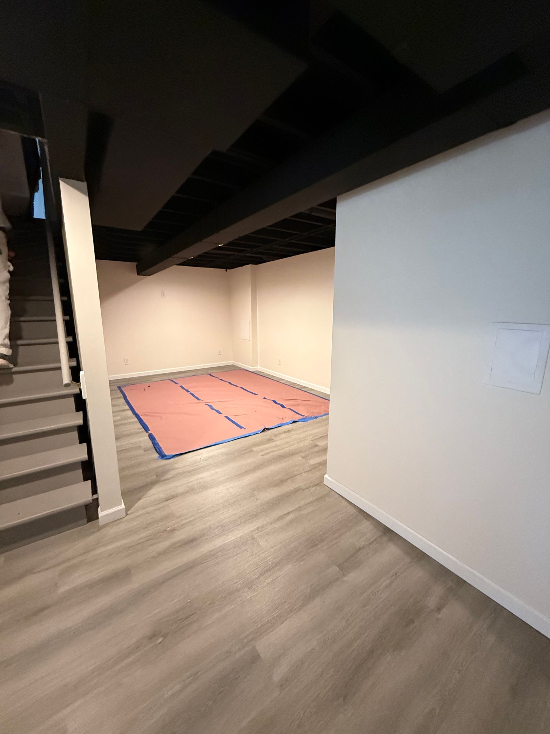 Basement room with gray flooring, black ceiling beams, white walls, and stairs. A mat covers the floor.
