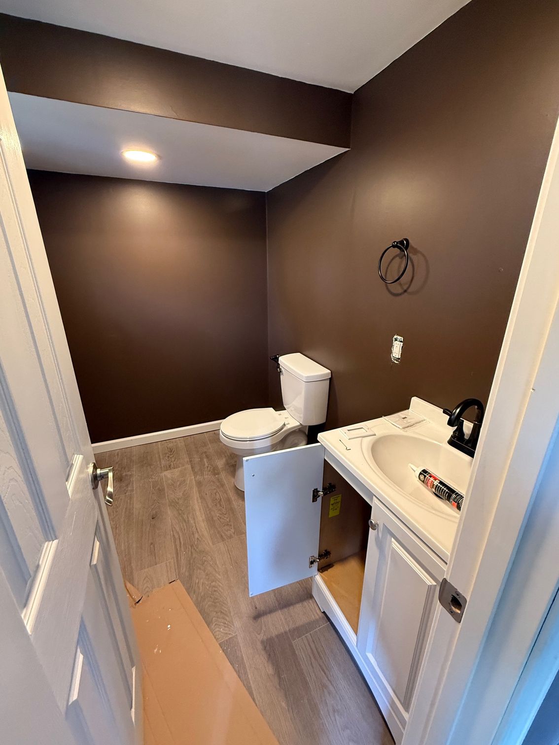 Small bathroom with brown walls, white toilet and sink, and light wood-look flooring.