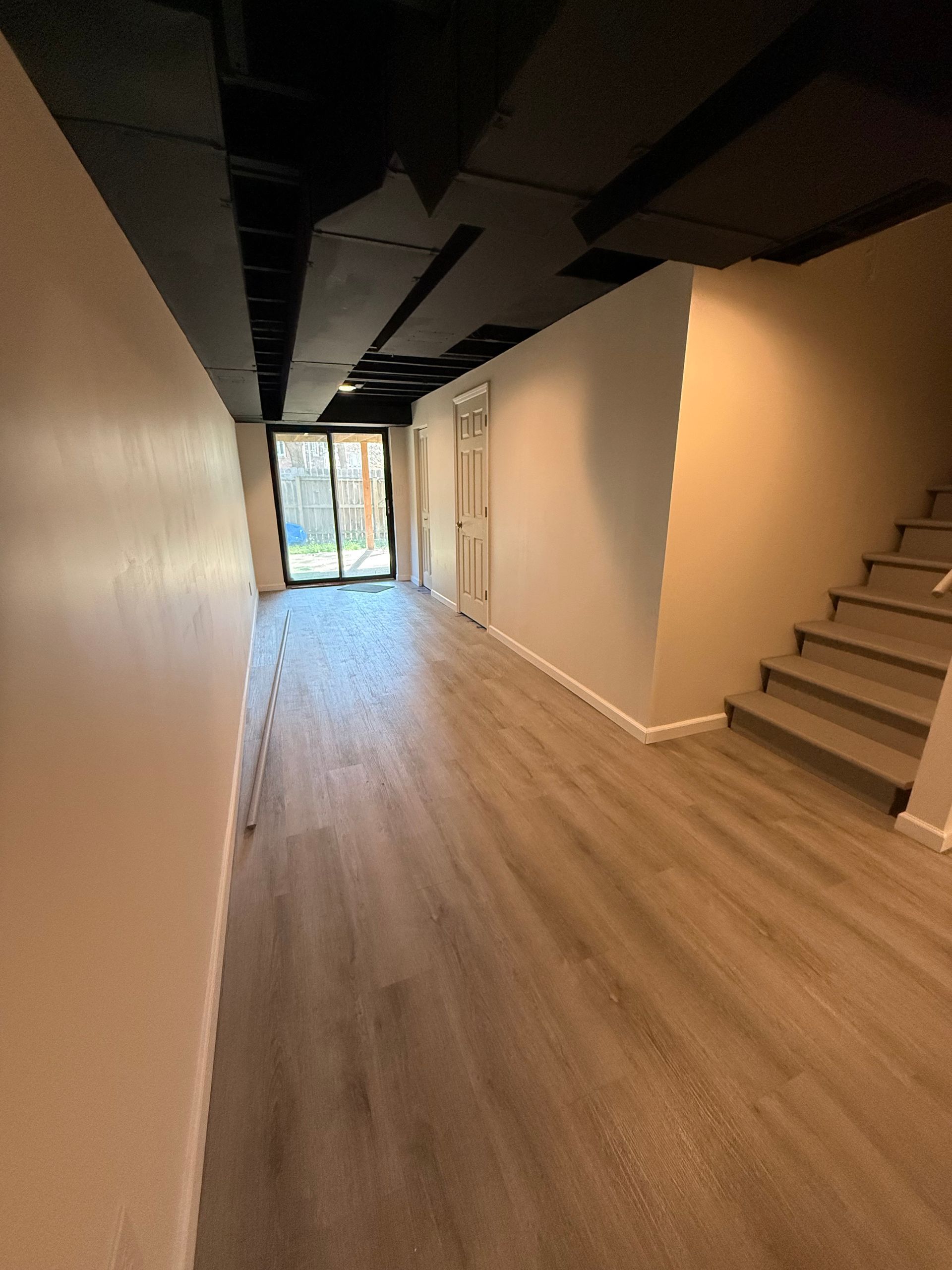 Basement hallway with light wood-look flooring, light-colored walls, and a black ceiling. Sliding glass door at the end.