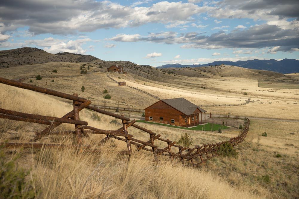 A log cabin in the middle of a dry grassy field with mountains in the background.