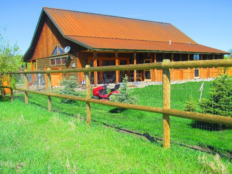 A red lawn mower is parked in front of a large barn behind a wooden fence.