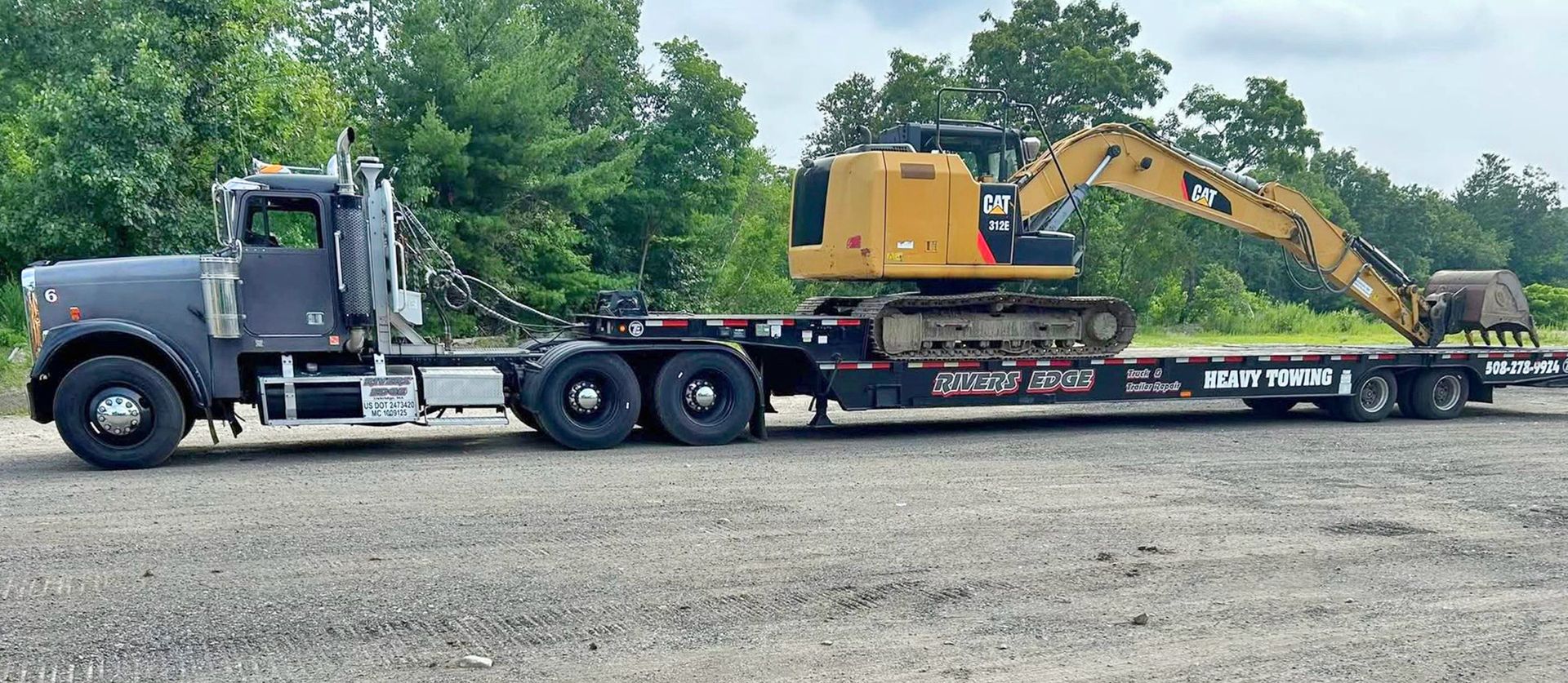 Truck hauling a yellow excavator on a flatbed trailer, parked on a gravel lot near trees.