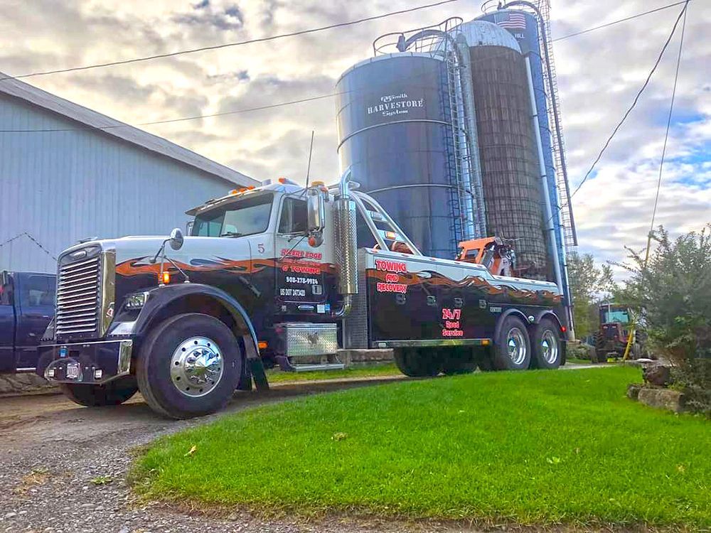 Tow truck parked next to a silo. The truck is black, white, and orange, with a logo and the sky is overcast.