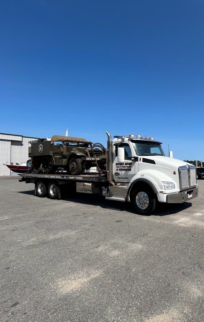 White tow truck transporting a military vehicle on a sunny day.