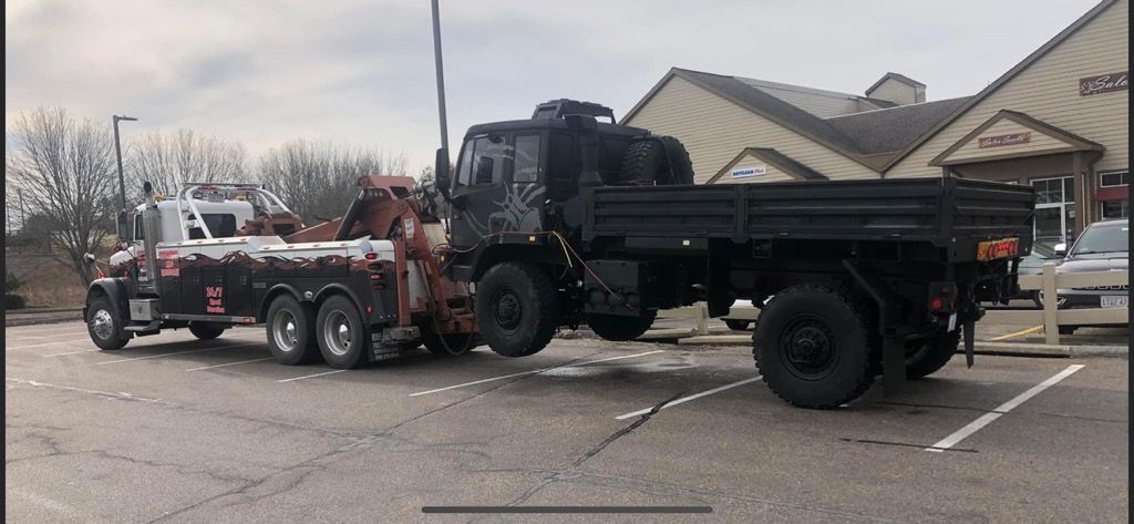 A black military truck is being towed by a tow truck in a parking lot.