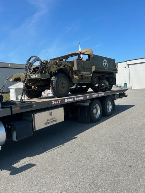 A military half-track vehicle on a flatbed tow truck. Green, weathered, under a blue sky.