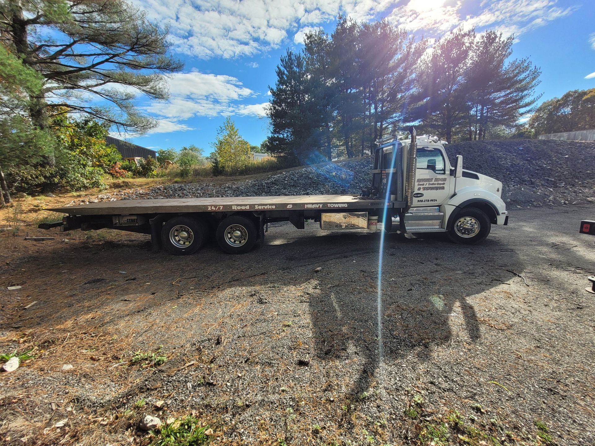White flatbed truck on gravel, outdoors. Sunny day, blue sky with clouds, trees in background.