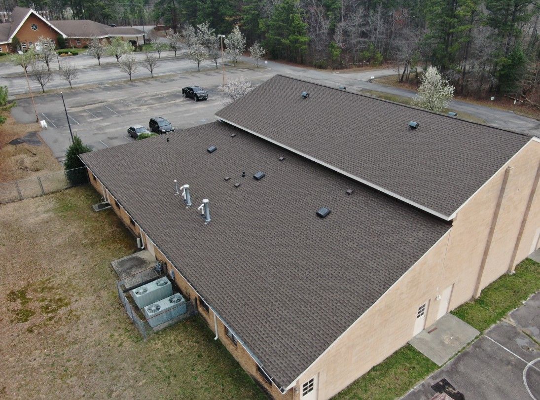 An aerial view of a brown-shingled roof on a tan brick building. Parking lot and trees in the background.