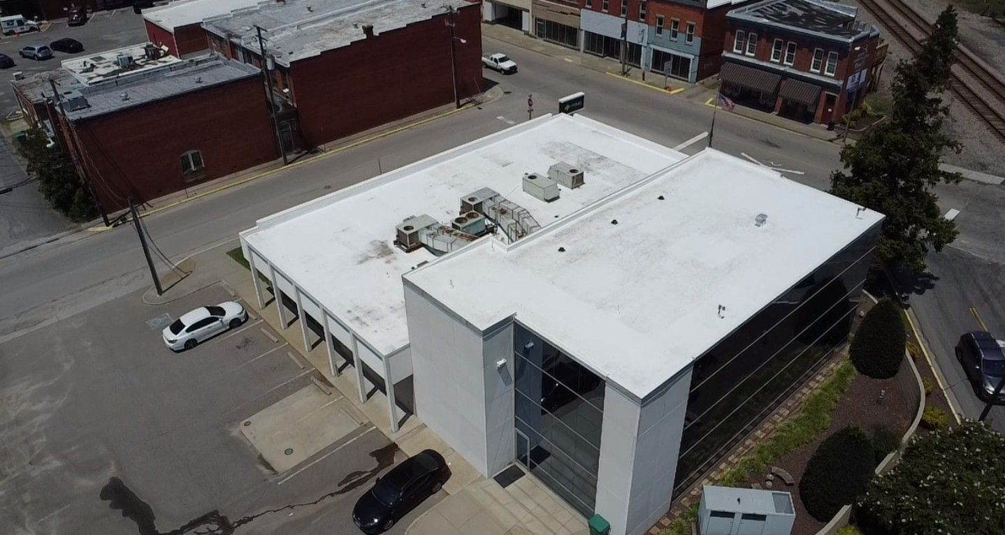 An aerial view of a two-story modern building with a flat white roof. Cars drive on nearby streets.