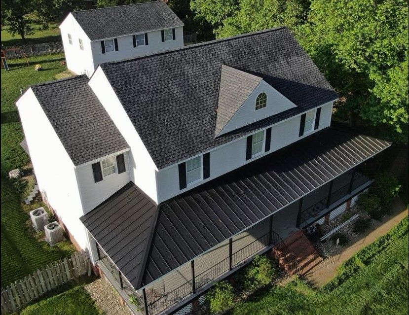 Aerial view of a white two-story house with black shingle roof, attached black metal porch, and separate white outbuilding.