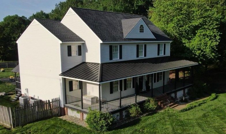 Two-story white house with a black roof and wrap-around porch, set against a backdrop of green trees.