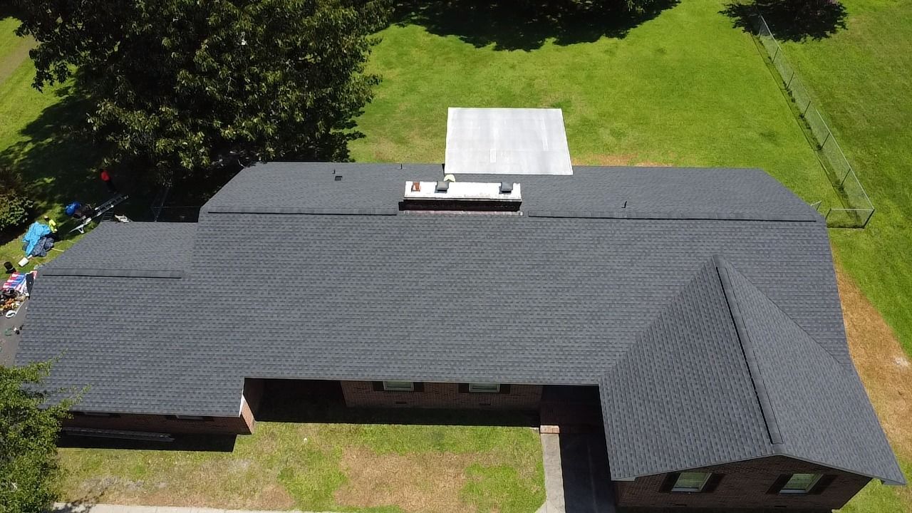 Aerial view of a dark gray shingled roof with a white rectangular structure and green grass surrounding.