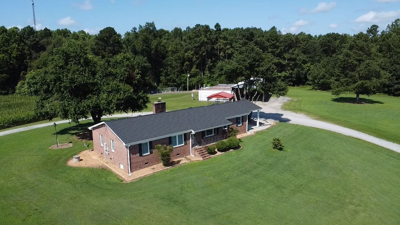Brick house with dark roof and chimney, surrounded by green grass, trees, and driveway on a sunny day.