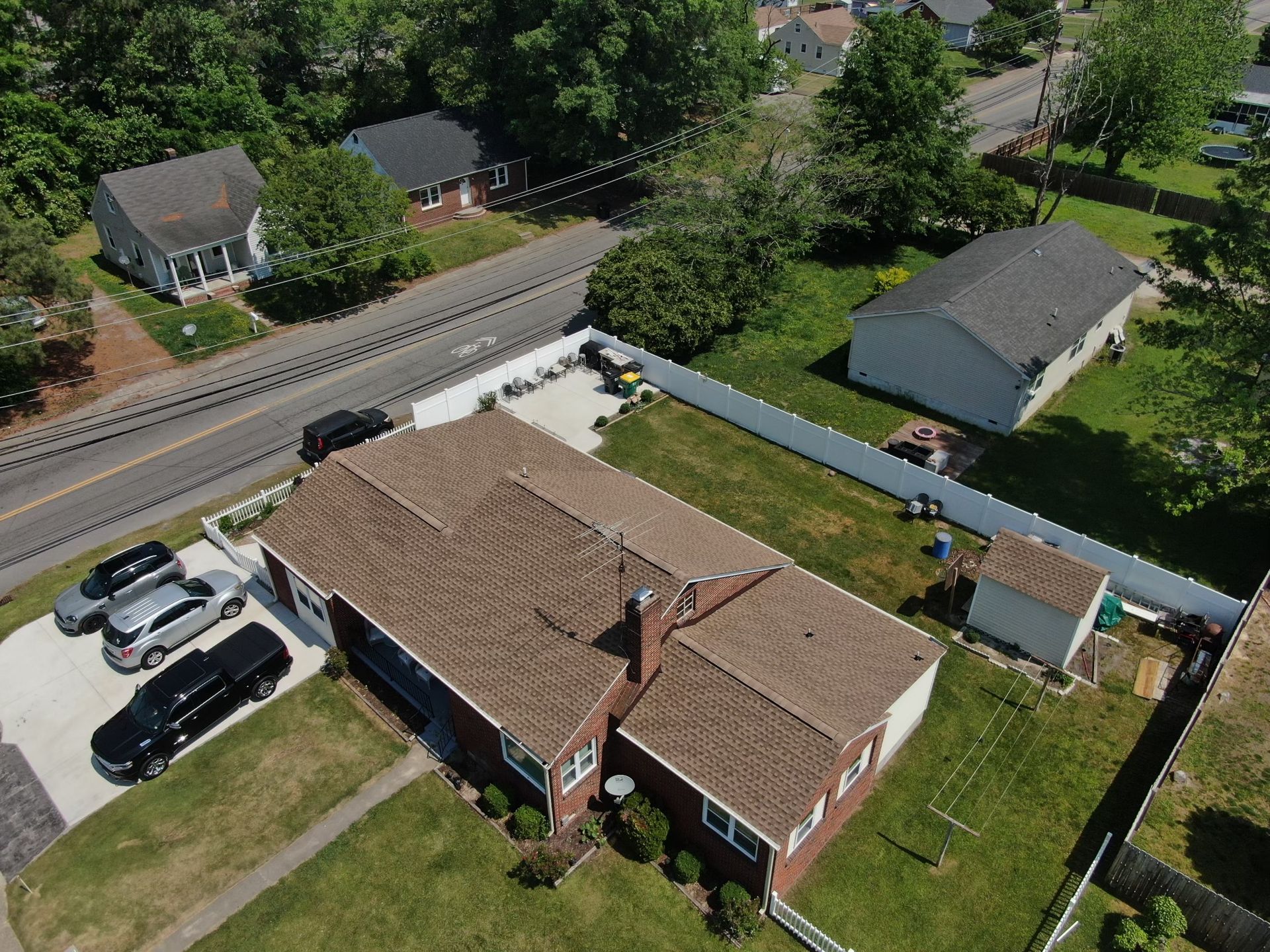 An aerial view of a house with a brown roof and a white fence. Cars are parked in the driveway.