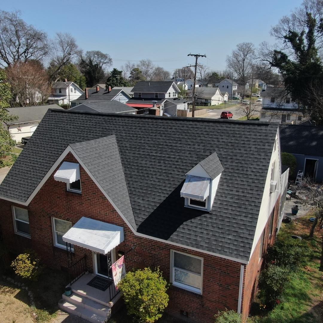 Brick house with dark roof and awnings, trees and other houses in background on a sunny day.