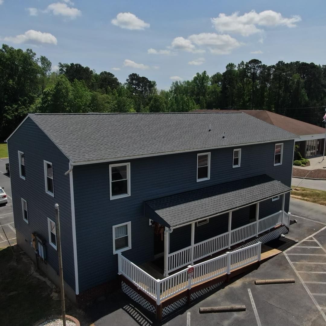 Two-story blue building with porch and dark roof, set in a parking lot with trees in the background.