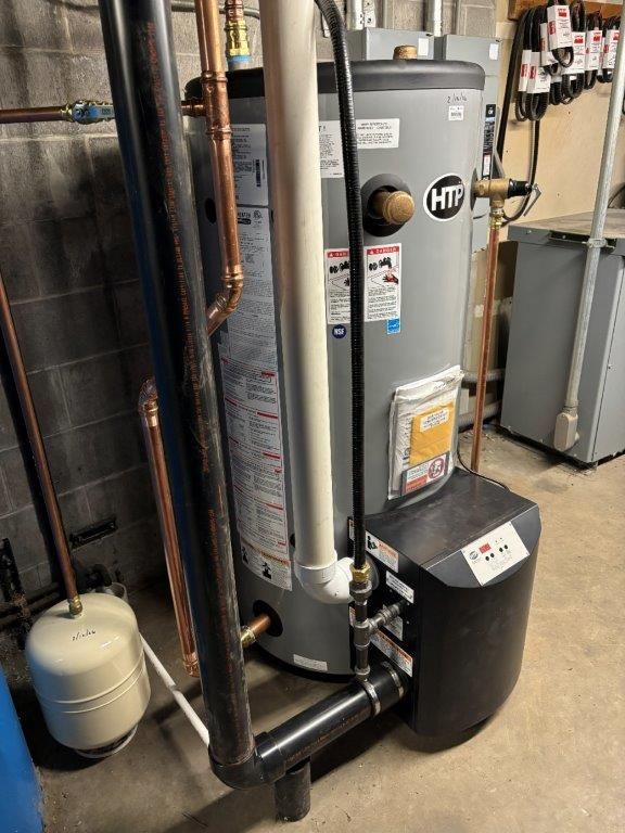 A tall, gray HTI water heater in a basement setting, with black pipes, copper piping, and a white pressure relief pipe.