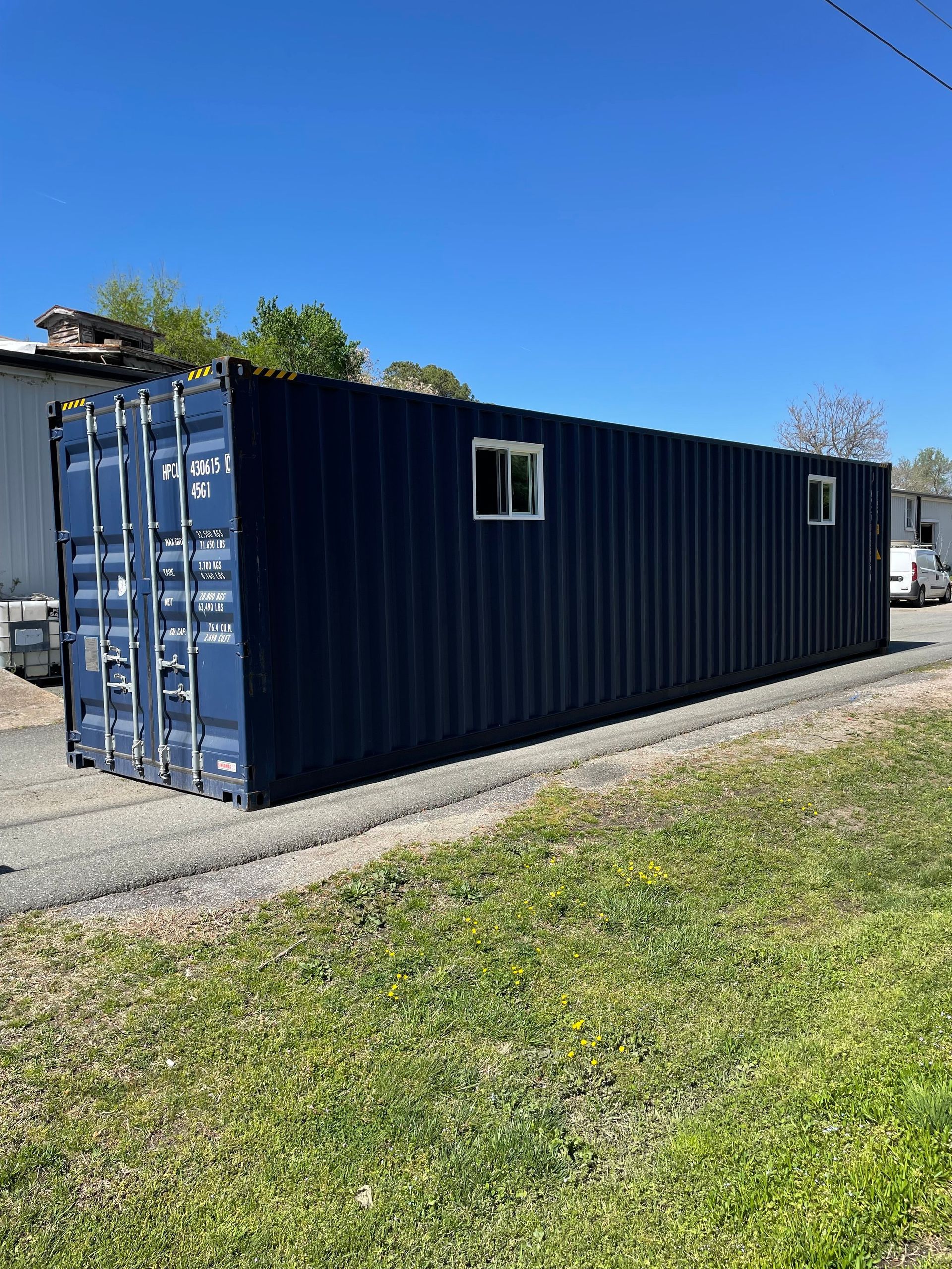 A black shipping container is sitting in the grass next to a road.