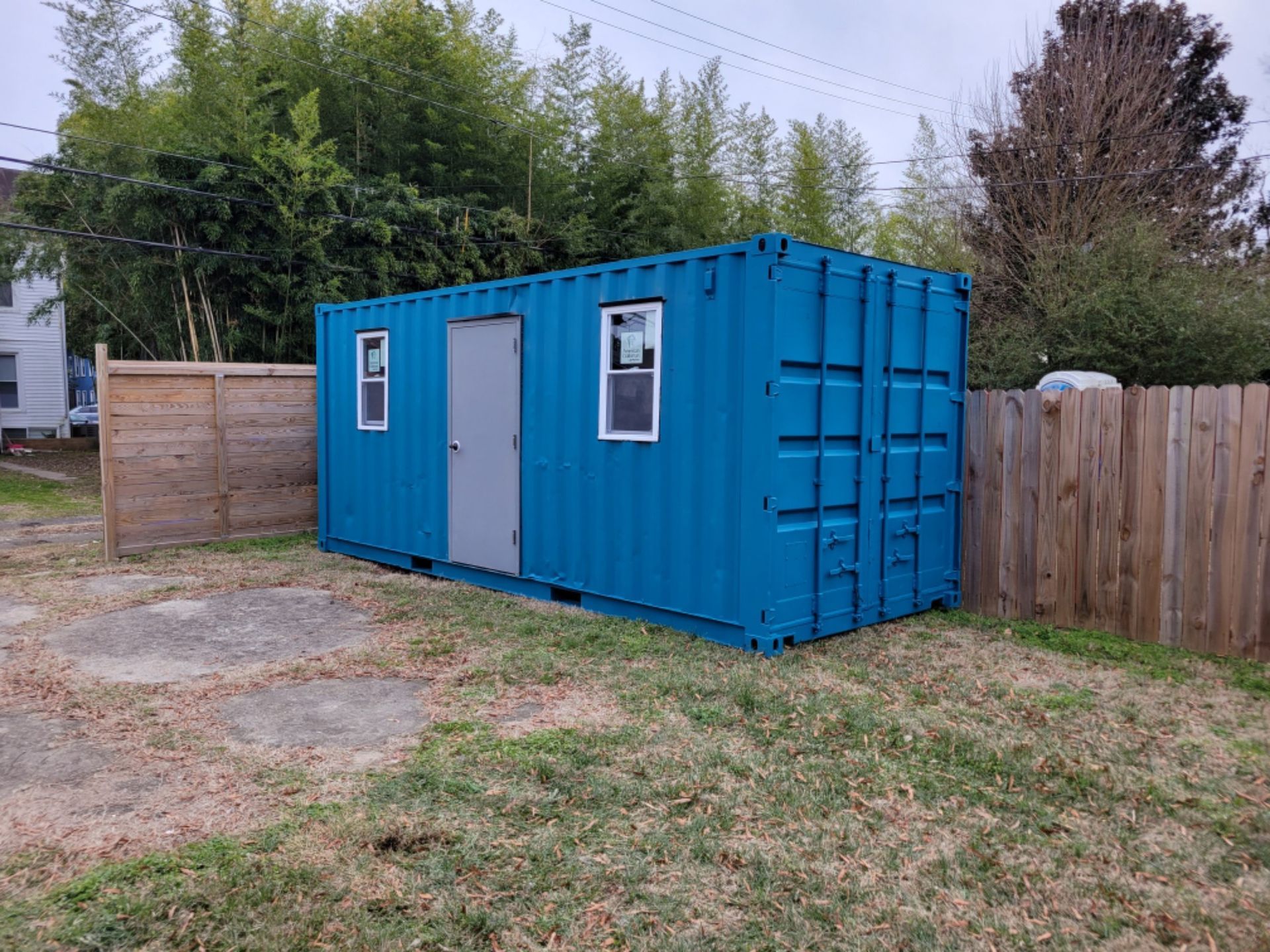 A blue shipping container with a door and windows is sitting in the grass next to a wooden fence.